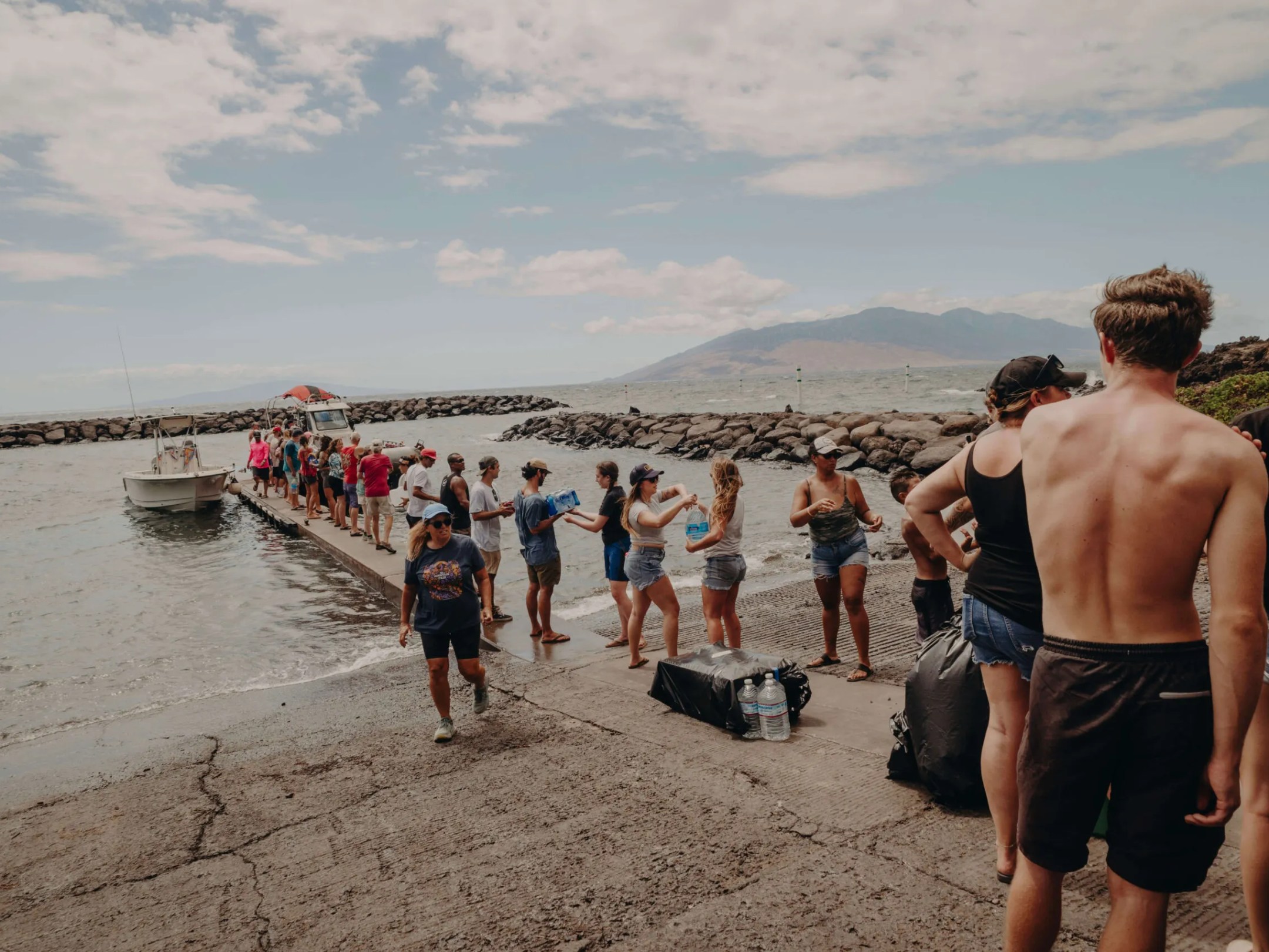 Group of people on a pier by the sea, passing supplies, with mountains in the background.