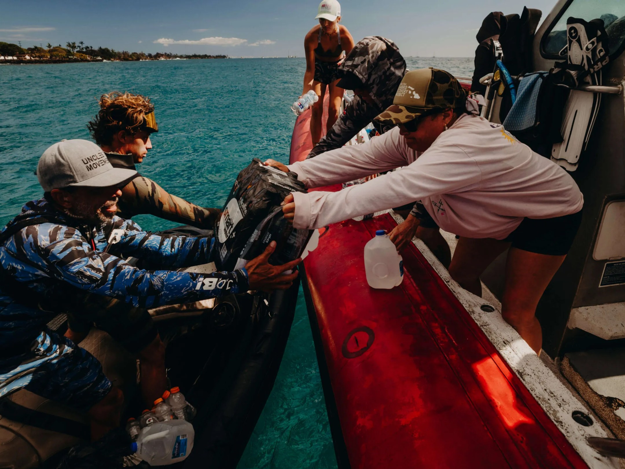 People on two boats passing supplies over clear blue water with coastline in the background.