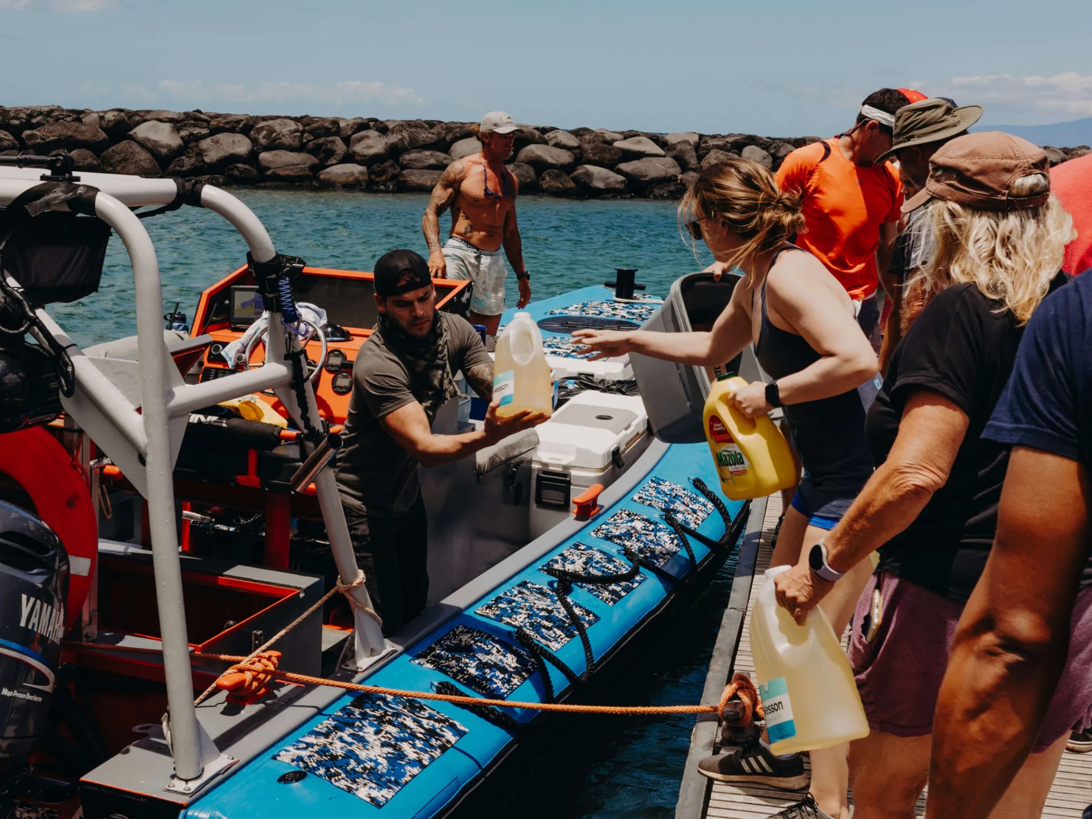 People load supplies onto a small boat at a dock under a clear sky.