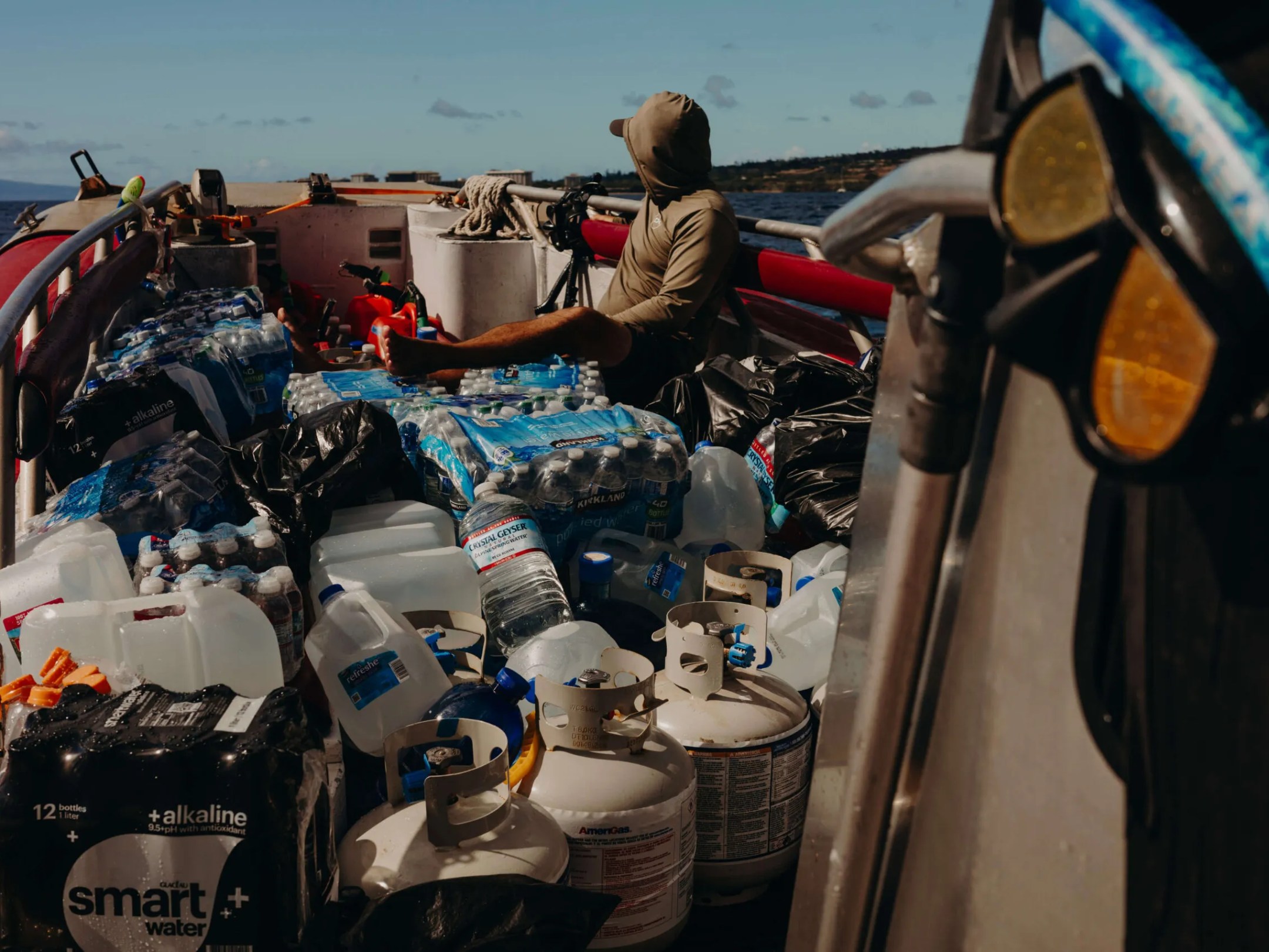 Person on a boat surrounded by water bottles and propane tanks under clear skies.