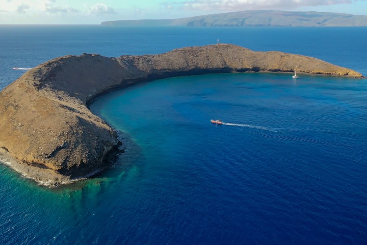 Aerial view of crescent-shaped island in blue ocean with boats nearby.