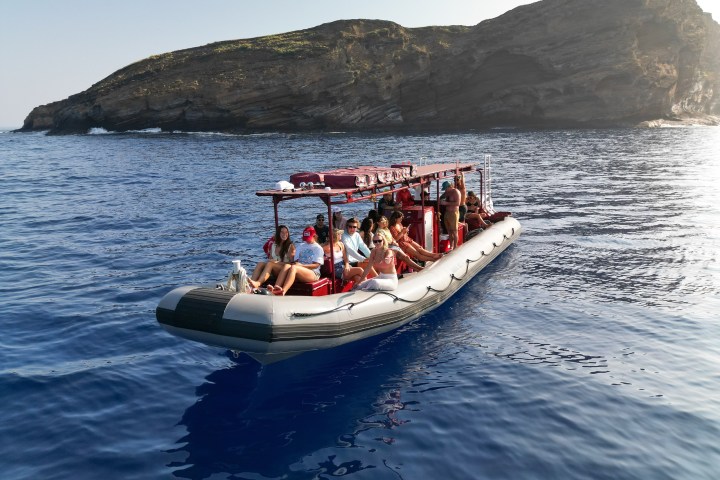 People on a small inflatable boat near a rocky island in clear blue water.