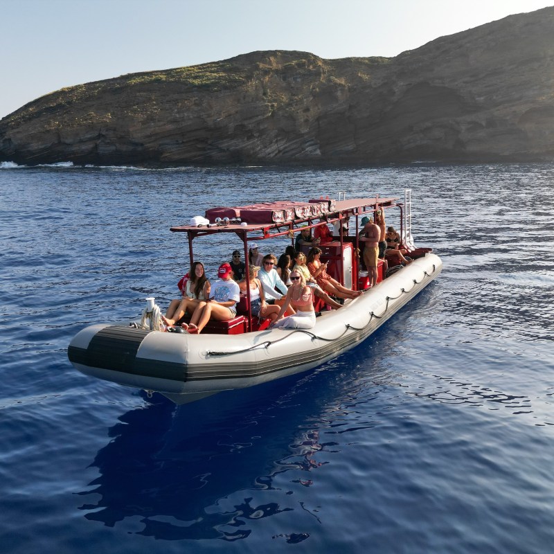 People on a small inflatable boat near a rocky island in clear blue water.