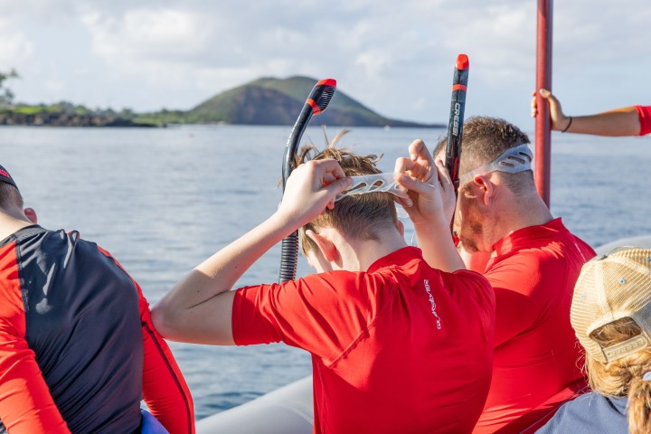 People in red shirts putting on snorkel masks on a boat near an island.
