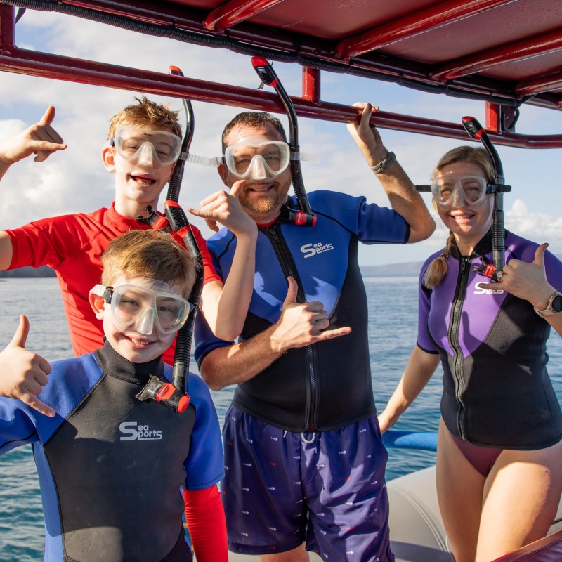 Four people in snorkeling gear on a boat, giving shaka signs, with water and sky in the background.