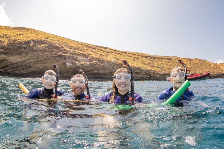 Four people wearing snorkeling gear smiling in the ocean near a rocky shoreline.
