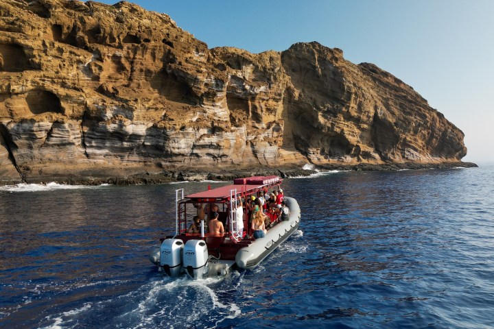 A boat with people approaches a large rocky cliff over blue ocean water under a clear sky.
