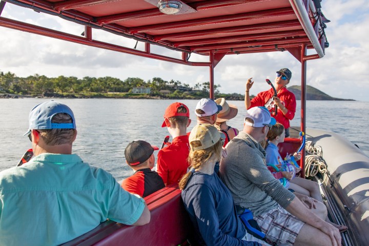 People on a boat with a guide speaking, surrounded by water and distant green landscape.