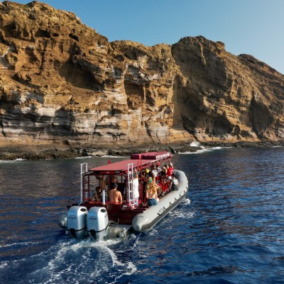 A boat with tourists approaches towering rocky cliffs over a calm blue ocean.