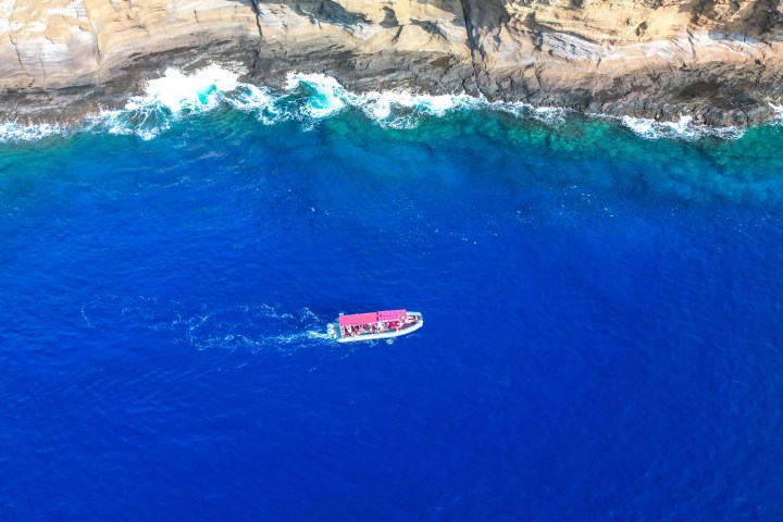 Aerial view of a boat with a red roof on deep blue ocean near rocky cliffs.