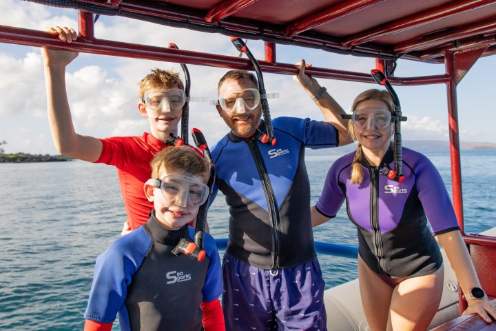 Four people in wetsuits and snorkel gear on a boat near the ocean.