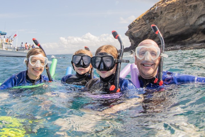 Four people snorkeling with swim noodles near a rocky island and a boat.