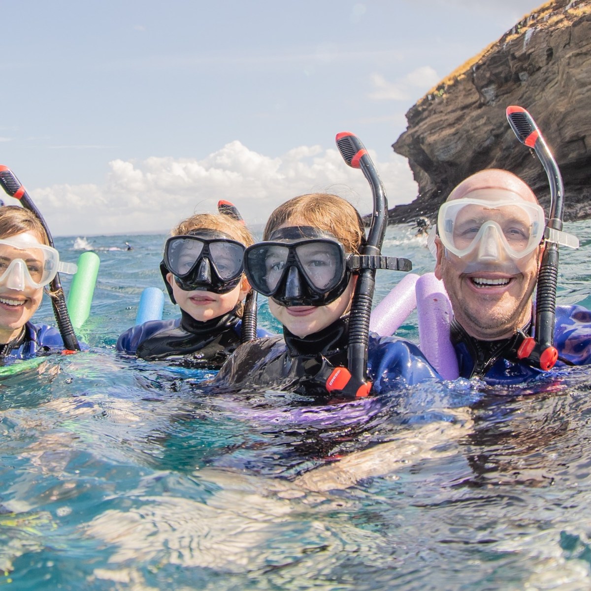 Four people snorkeling with swim noodles near a rocky island and a boat.