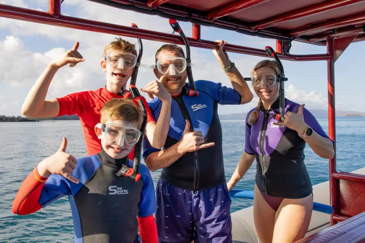 Four people in snorkeling gear on a boat, giving shaka signs, with water and sky in the background.