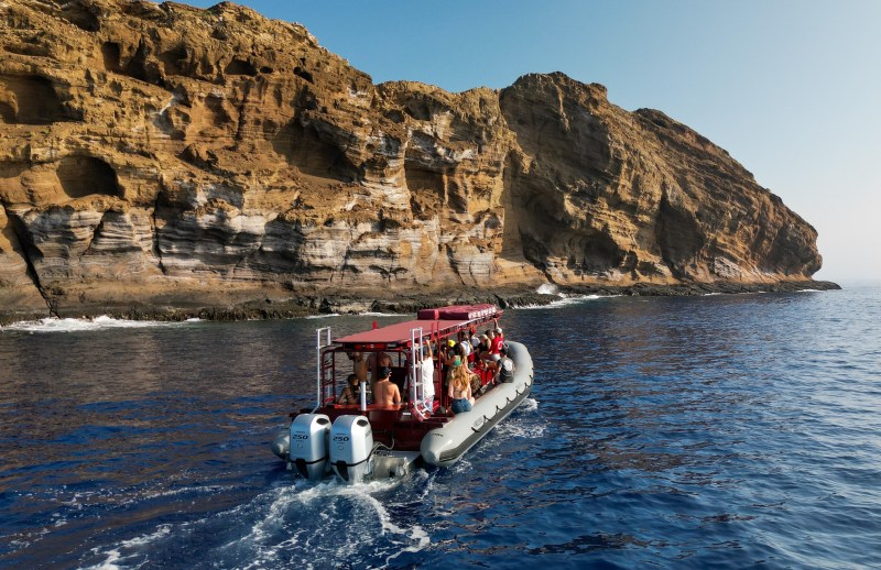 A boat with people approaches a large rocky cliff over blue ocean water under a clear sky.